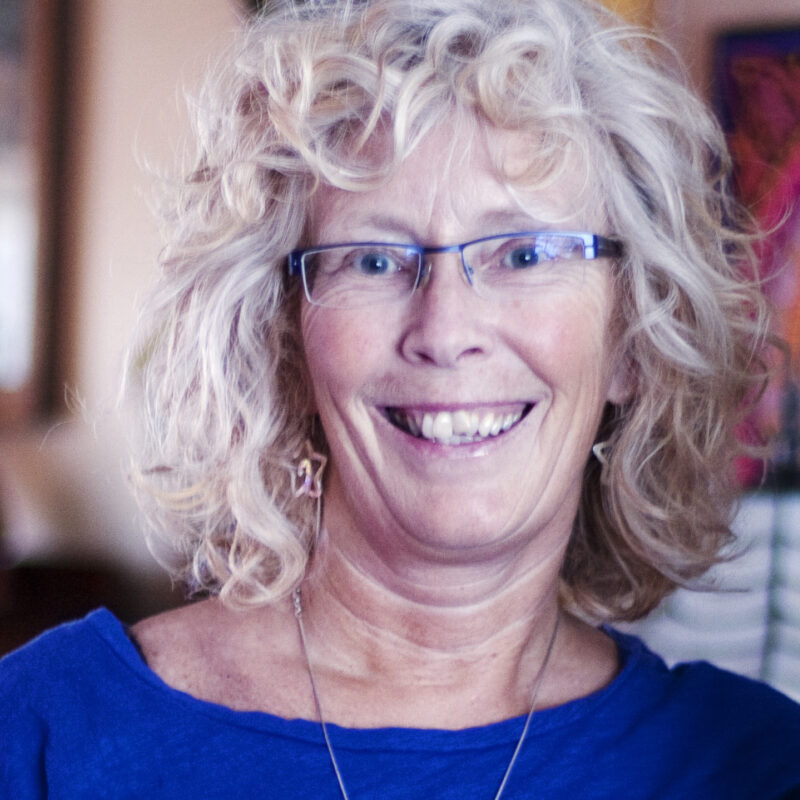 Person with curly gray hair and glasses wearing a blue shirt, smiling at the camera indoors.