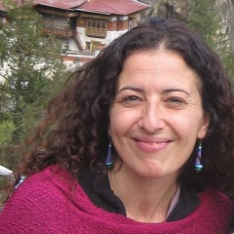 Person with long dark curly hair wearing a pink/magenta sweater and blue earrings, smiling at camera outdoors with house visible behind.