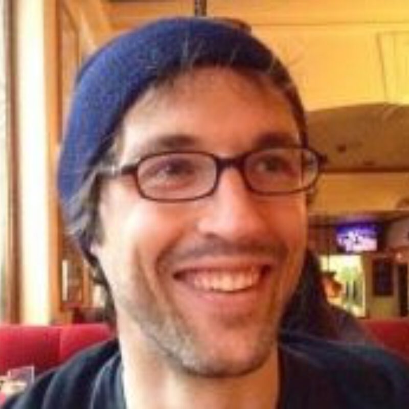 Person wearing glasses and a dark blue beanie, smiling at the camera indoors.