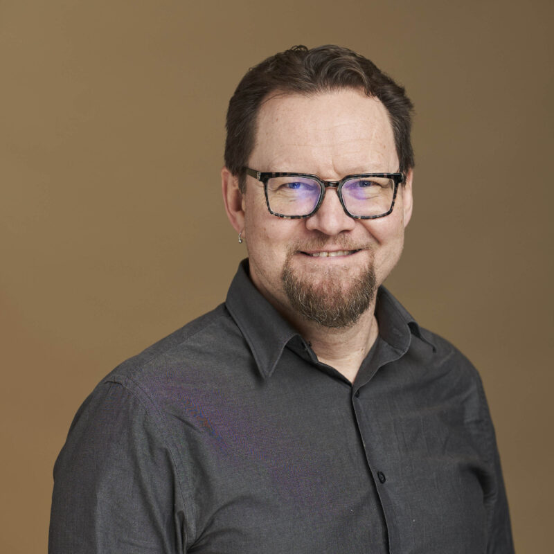 Person with glasses and beard wearing dark button-up shirt against tan background, smiling at camera.