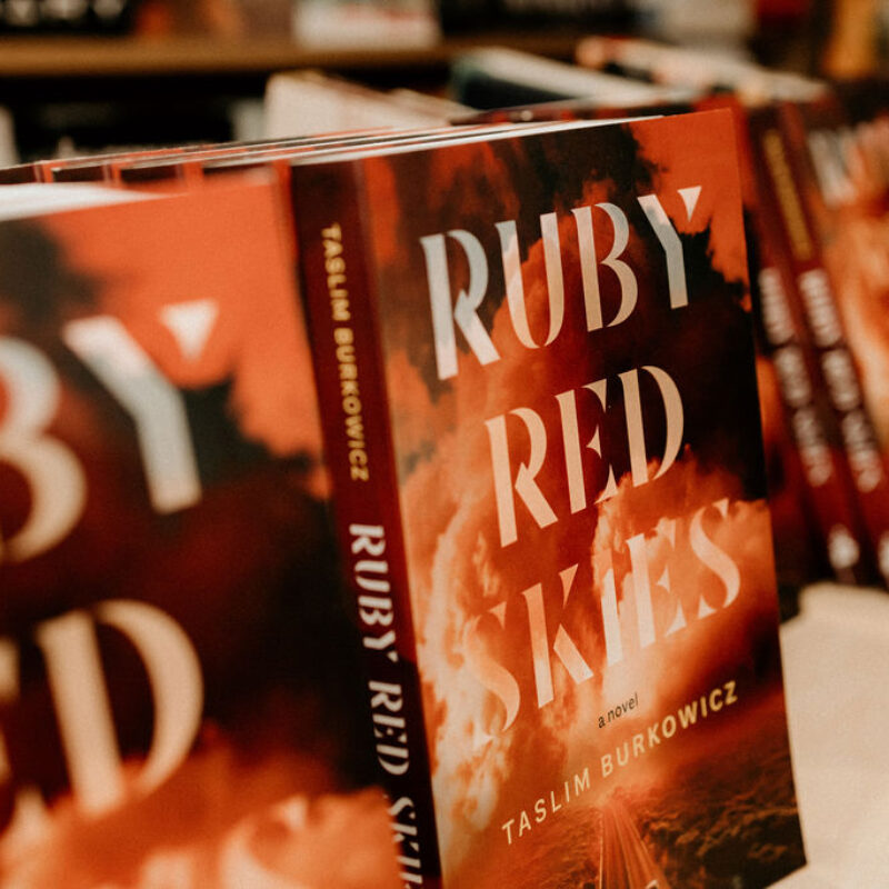 Multiple copies of "Ruby Red Skies" by Taslim Burkowicz displayed on a shelf with orange book covers.
