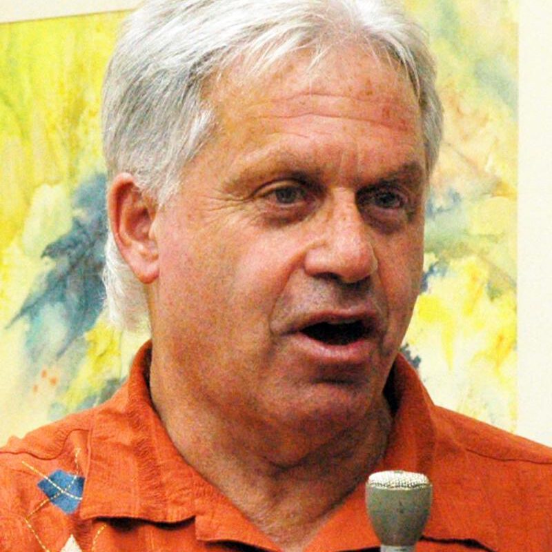 An older person with white hair wearing a bright orange shirt speaks into a microphone in front of a yellow-green watercolor painting.