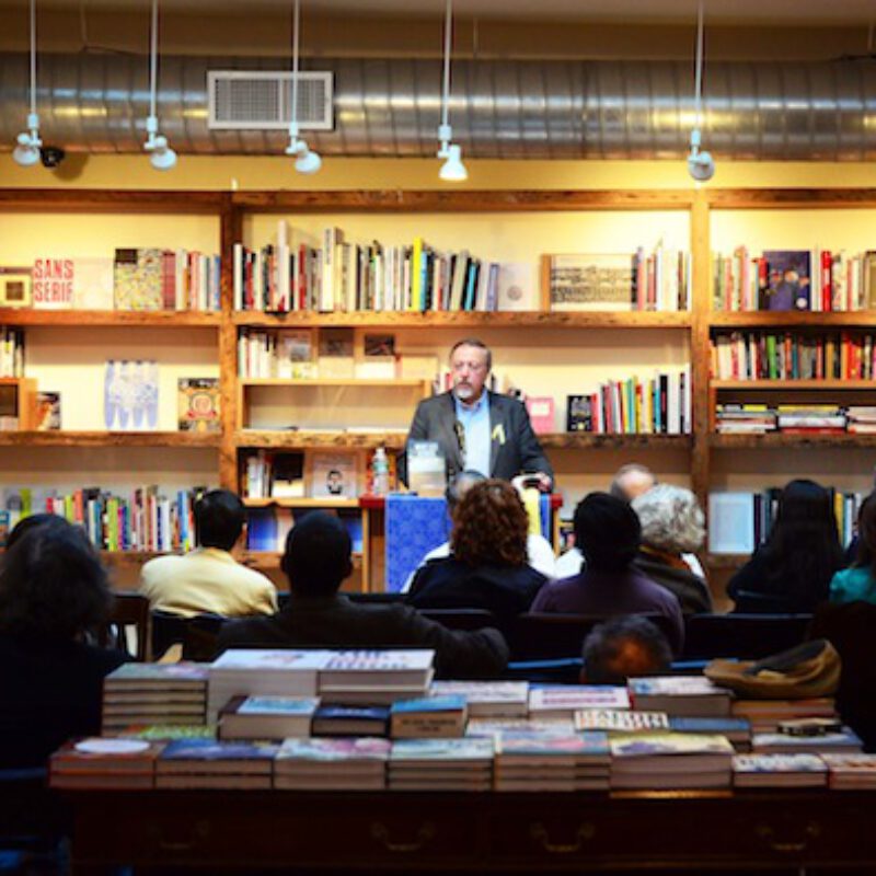 Person speaking at podium in bookstore with shelves of books behind and audience seated in front.