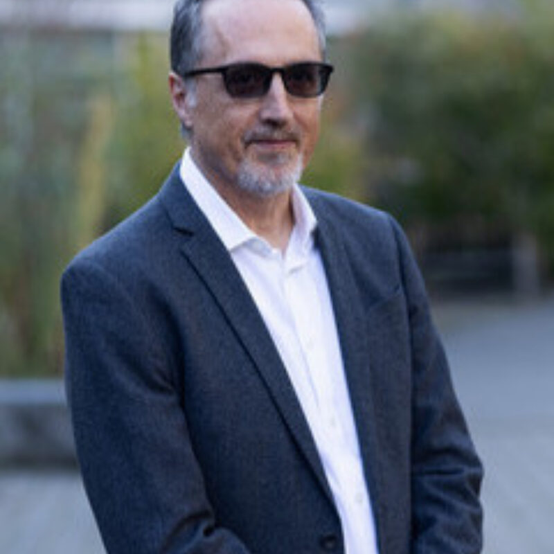 Person wearing dark blazer and white shirt with sunglasses standing outdoors on a street.