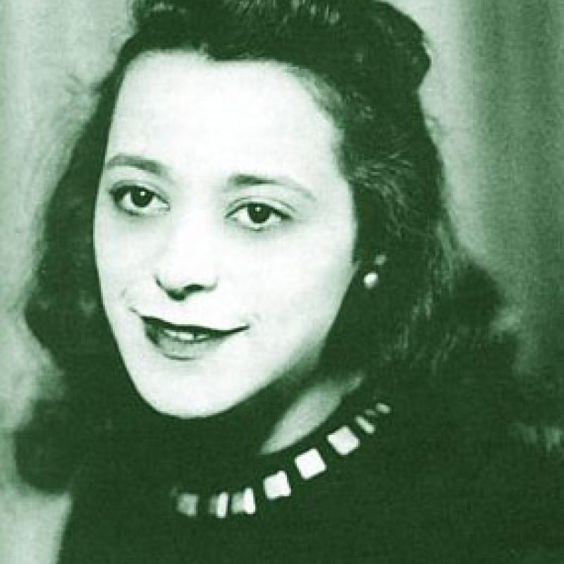 Black and white portrait photograph of a person wearing a dark top with decorative studded neckline, pearl earrings, and styled dark hair.