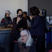 Group of people gathered in an indoor space, some standing by a counter and others seated, appearing to be in conversation or at a social event.