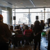People gathered in a high-rise room with city views, seated and standing around tables during what appears to be a community or educational event.