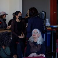 People gather indoors at what appears to be a community event or meeting, with some standing and conversing while others sit in chairs.
