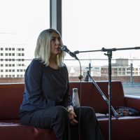 Person with blonde hair sitting on red couch speaking at microphone with city waterfront view behind.
