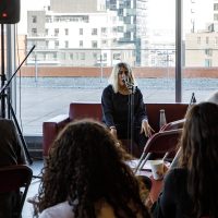 Speaker at microphone addressing audience in modern venue with waterfront and city buildings visible through windows.