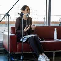 A person speaks at a microphone on a red couch in a high-rise venue overlooking a waterfront and city skyline.