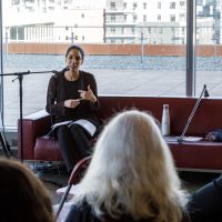 Speaker sitting on burgundy couch addresses audience during indoor event with city skyline visible through windows behind them.