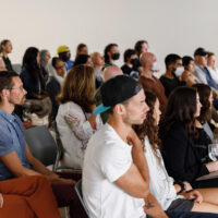 A diverse group of people sitting and standing in an auditorium, attentively watching a presentation or event.
