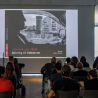 A presenter stands before a seated audience while a projection screen displays "Driving in Palestine" with Arabic text and architectural imagery.