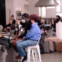 Two people sit on a white bar stool in a modern kitchen during a community gathering with multiple attendees seated around them.