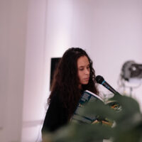 Person with long dark hair speaking at a microphone while holding a document in an indoor venue with white walls.