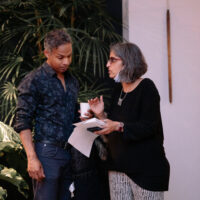 Two people having a conversation in front of a potted plant, one holding a cup and papers while wearing glasses.
