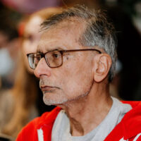 Older person wearing glasses and red jacket at an indoor event with blurred audience in background.