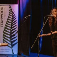 Person speaking at microphone on stage with Fernwood Publishing banner and blue lighting in background.