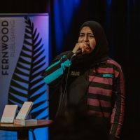 Person speaking at microphone on stage wearing hijab and striped jacket, with Fernwood Publishing backdrop and books on table.