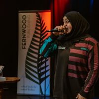 Person wearing hijab speaking into microphone at Fernwood Publishing event on stage.