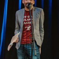 Person at microphone on stage wearing gray blazer and red graphic t-shirt, holding papers, with blue lighting backdrop.