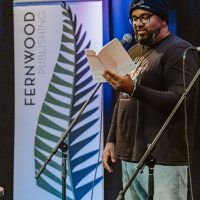 Person reading from a book at a microphone during a Fernwood Publishing event, with blue stage lighting in the background.