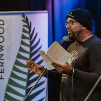 A person wearing glasses and a beanie holds an open book while speaking at a microphone, with a Fernwood Publishing banner visible behind them.
