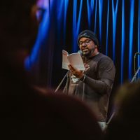 Person wearing glasses and beanie reading from paper at microphone on stage with blue lighting and audience in foreground.