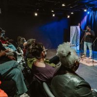 Audience members seated in theater watching two people on stage near a microphone and podium with blue lighting.