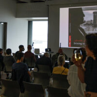 Audience members at an indoor presentation watching a projected image on a screen in a modern venue.